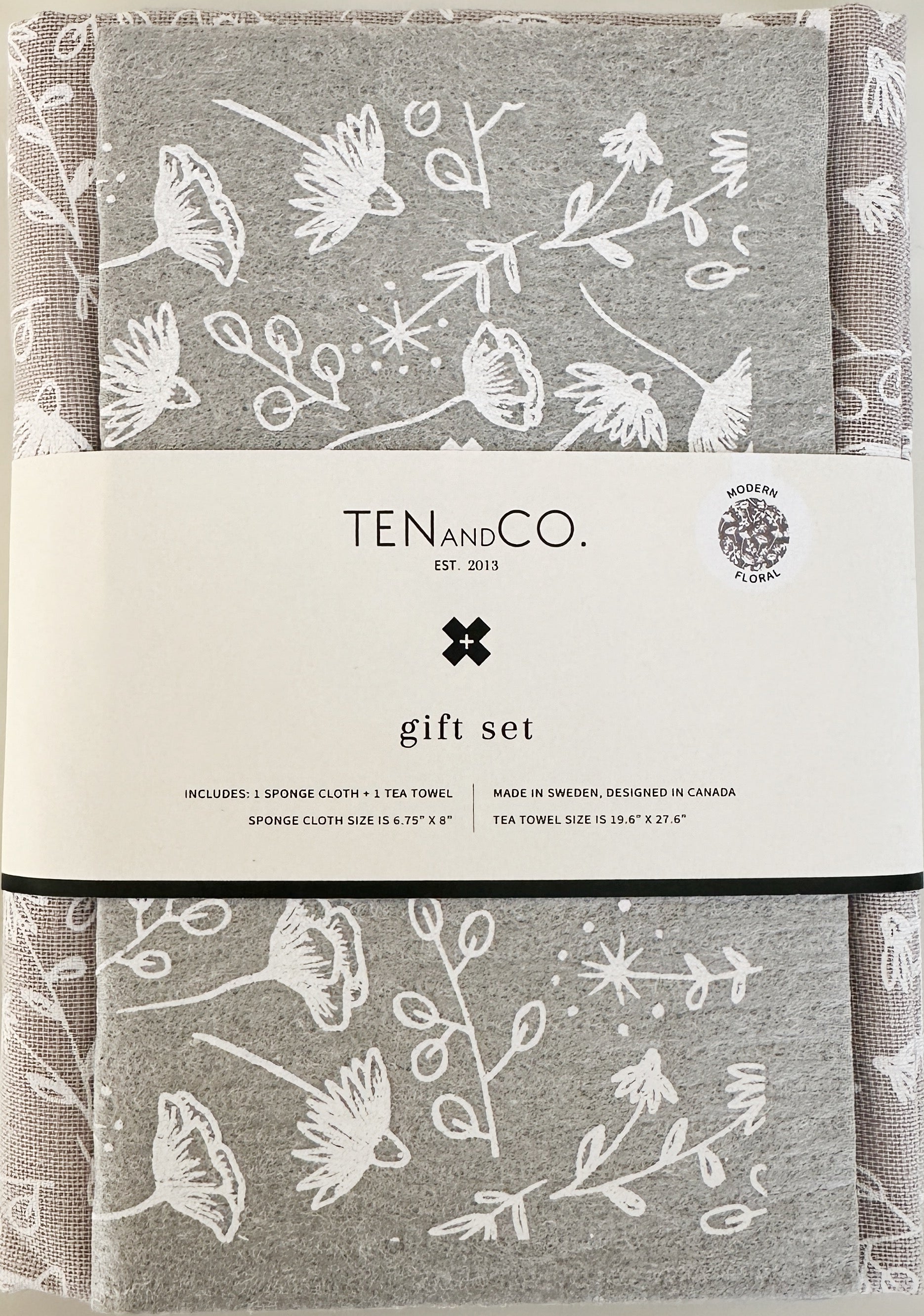 Flat lay image of Modern Floral gift set. There is a Modern Floral sponge cloth folded with a Modern Floral tea towel behind it. There is a white belly band across both. The sponge cloth has a grey base with white flowers throughout. The tea towel has a white base with black flowers throughout. The belly band has the Ten and Co. Logo and descriptive text in black and grey font.