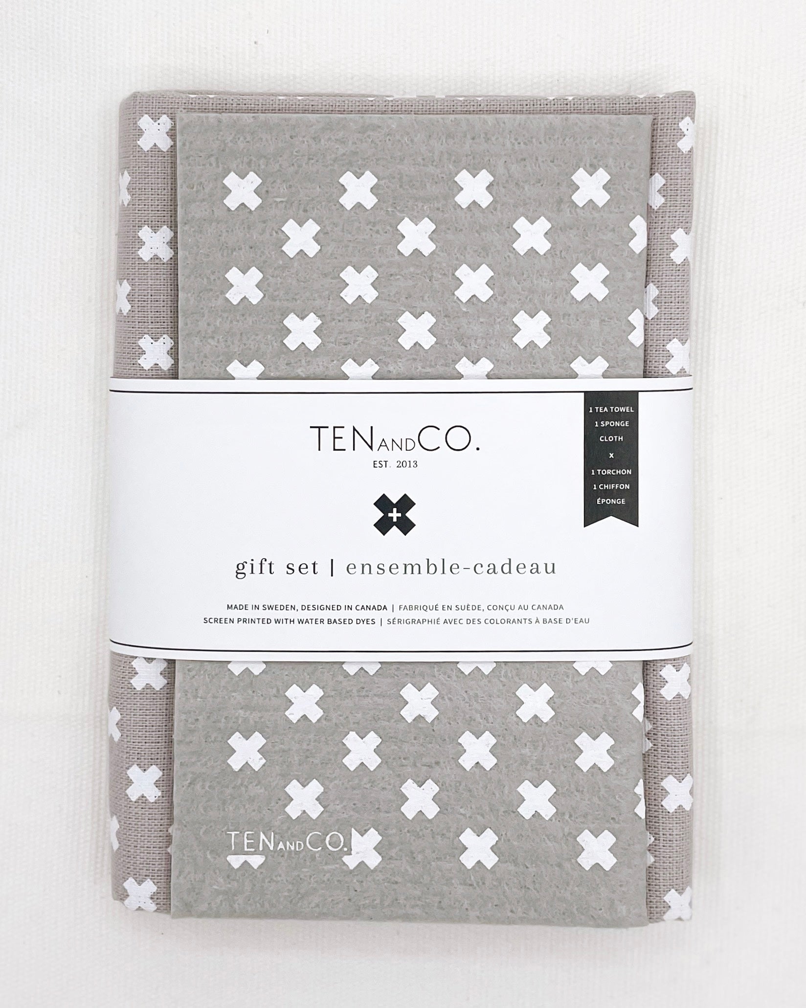 Flat lay image of Tiny X White on Warm Grey gift set on a white background. There is a Tiny X White on Warm Grey sponge cloth folded with a Tiny X White on Warm Grey tea towel behind it. There is a white belly band across both. The sponge cloth has a grey base with tiny x’s in white throughout. The tea towel has a grey base with tiny x’s in white throughout. The belly band has the Ten and Co. Logo and descriptive text in black font.